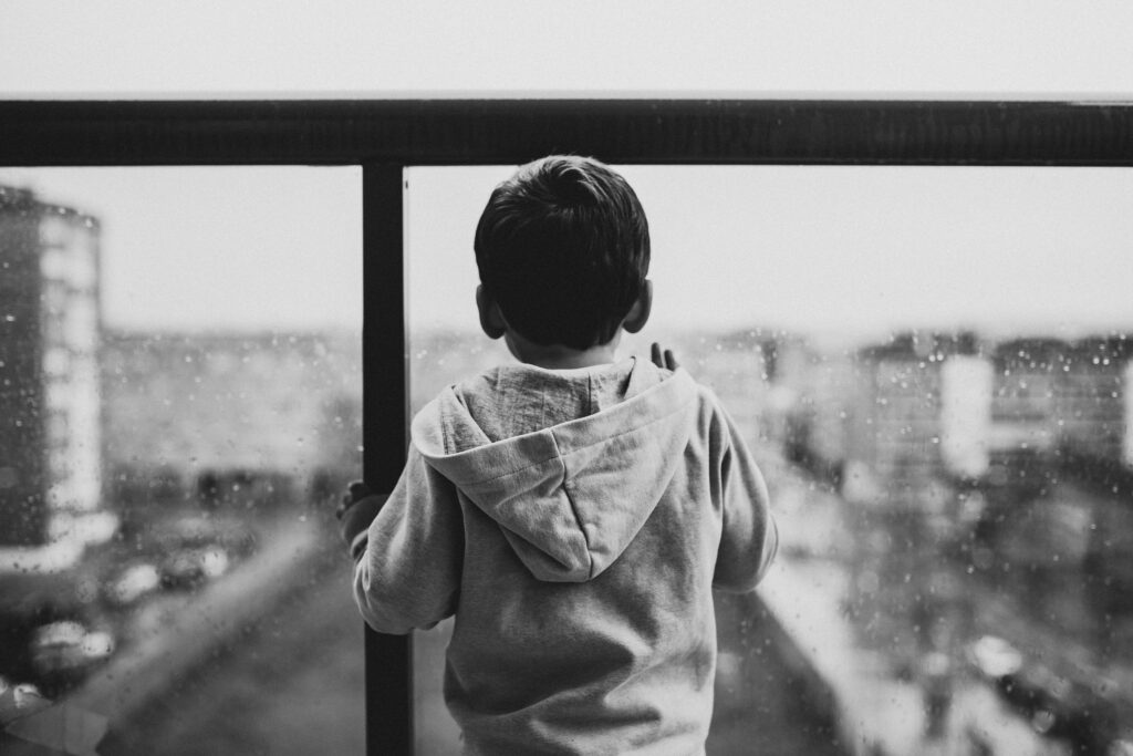 A young boy in a hoodie looks out a rainy window, creating a moody black-and-white scene.